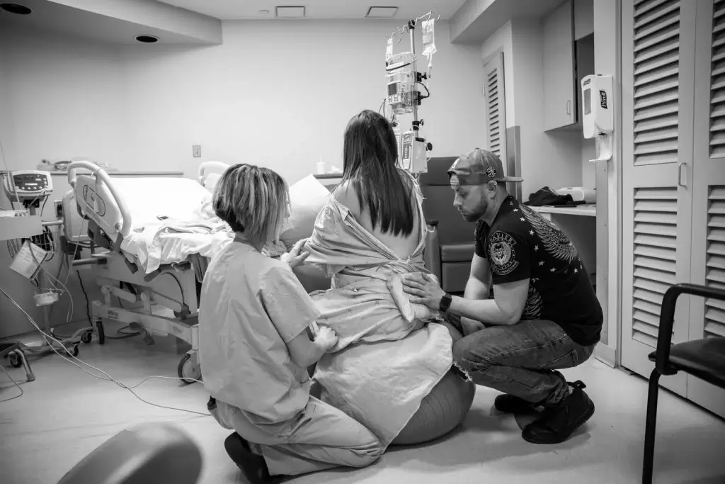 lady labouring on a birthing ball with a nurse and the partner supporting her. partner is holding a heat pack to her back
